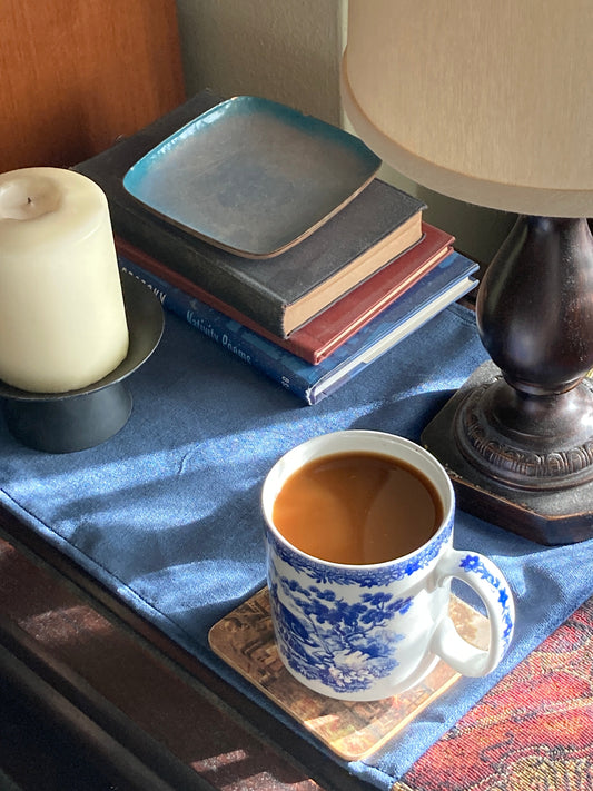 Coffee in a spode cup on a velvet table runner near a lamp, candle, and stack of books